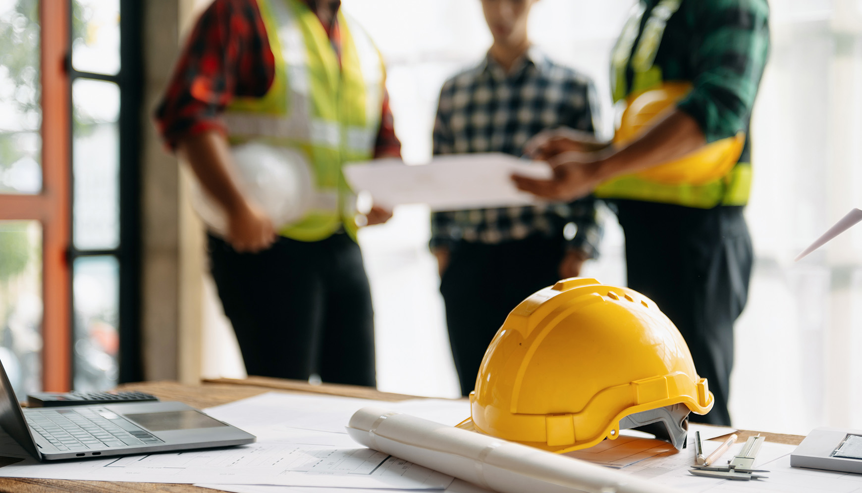 Construction workers discussing safety plans with a yellow hard hat and blueprints on a table, emphasizing workplace safety and OSHA compliance.