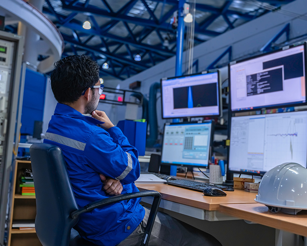 Man in blue safety jacket observing multiple computer monitors in a workplace setting, emphasizing safety and compliance in industrial operations.