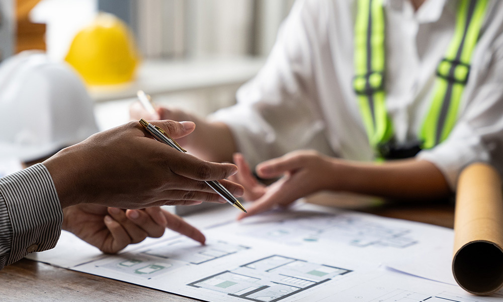Hands discussing construction plans on a table with safety helmets and blueprints, emphasizing OSHA compliance and inspection support services.
