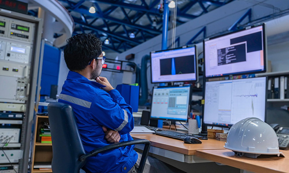 Engineer in blue safety jacket analyzing data on multiple computer screens in an industrial setting, with a white hard hat on the desk, emphasizing workplace safety and compliance.
