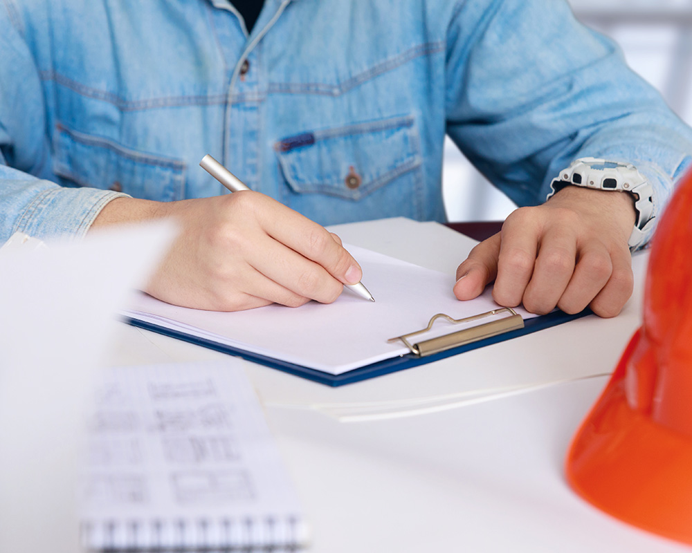 Person writing notes on clipboard with safety helmet and documents, emphasizing workplace safety and OSHA compliance resources.