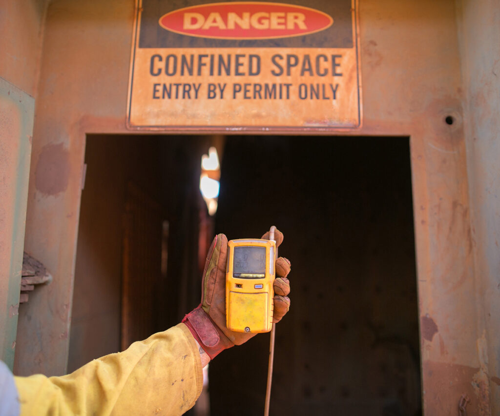 Worker holding gas detector in front of "Danger: Confined Space" sign, emphasizing safety measures for OSHA compliance in workplace environments.