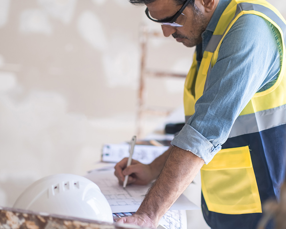 Construction worker in safety vest reviewing plans on a table, with a hard hat nearby, emphasizing workplace safety and OSHA compliance.