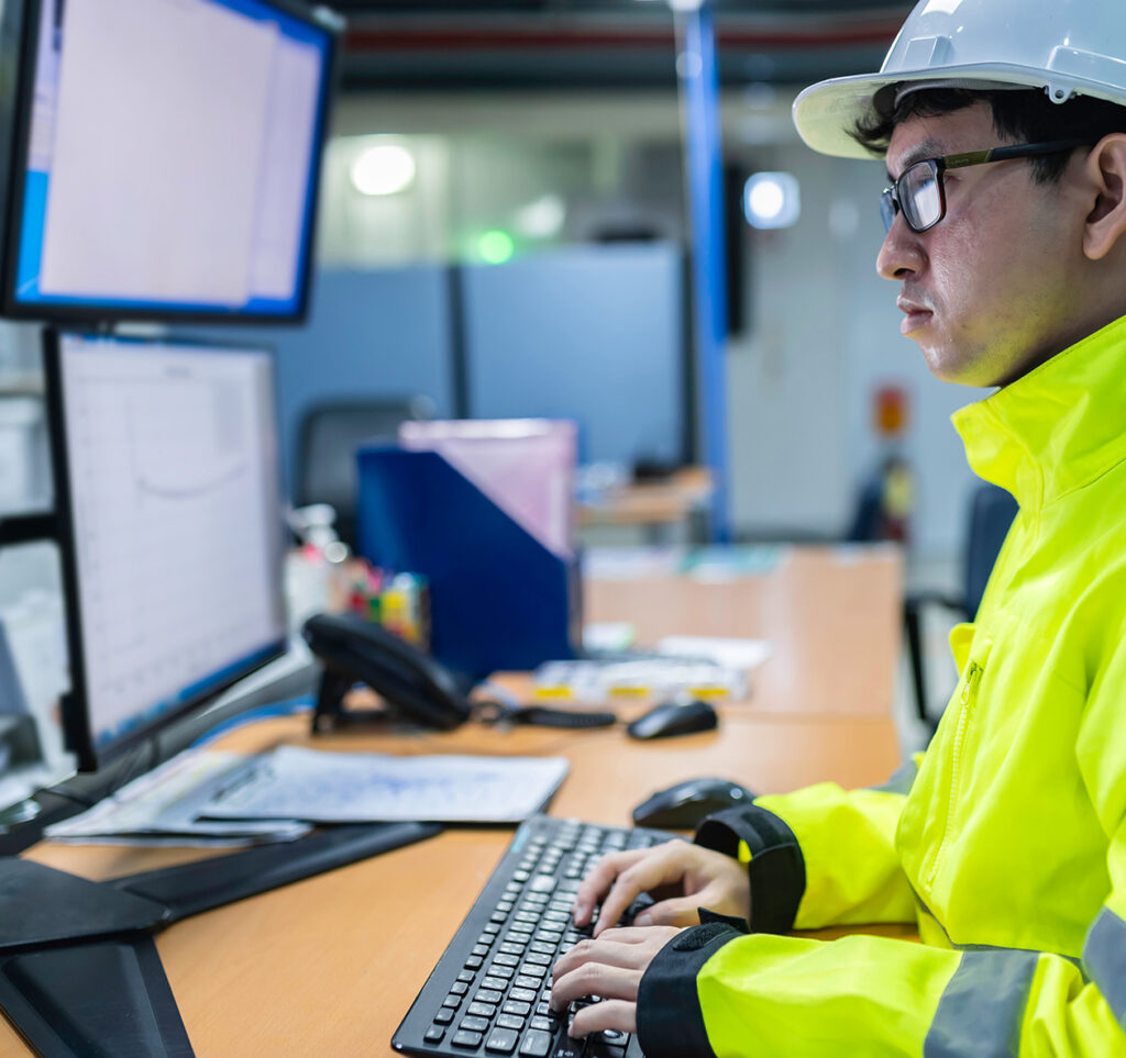 Person in safety gear typing on a keyboard at a workstation, with monitors displaying data, illustrating workplace safety program development and OSHA compliance.