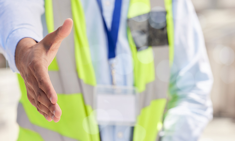 Construction professional extending hand for handshake, wearing safety vest and ID badge, symbolizing contractor vetting and compliance support in Phoenix area.