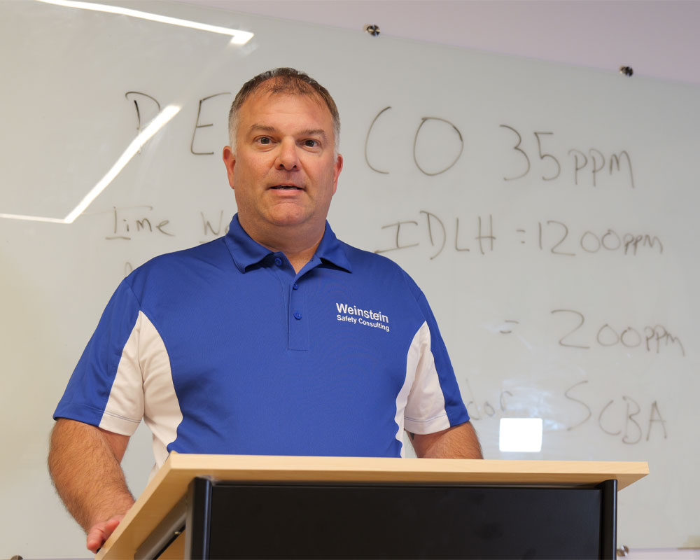 Man in blue and white shirt with "Weinstein Safety Consulting" logo, standing at podium in front of whiteboard displaying safety training information on exposure limits and equipment.