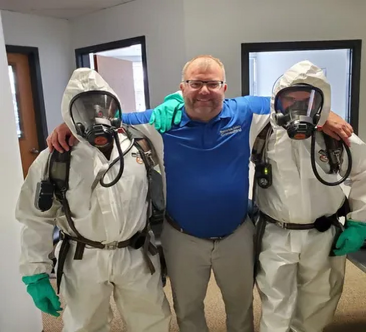 Man in blue shirt with two individuals in protective hazmat suits, emphasizing safety training and OSHA compliance in workplace environments.