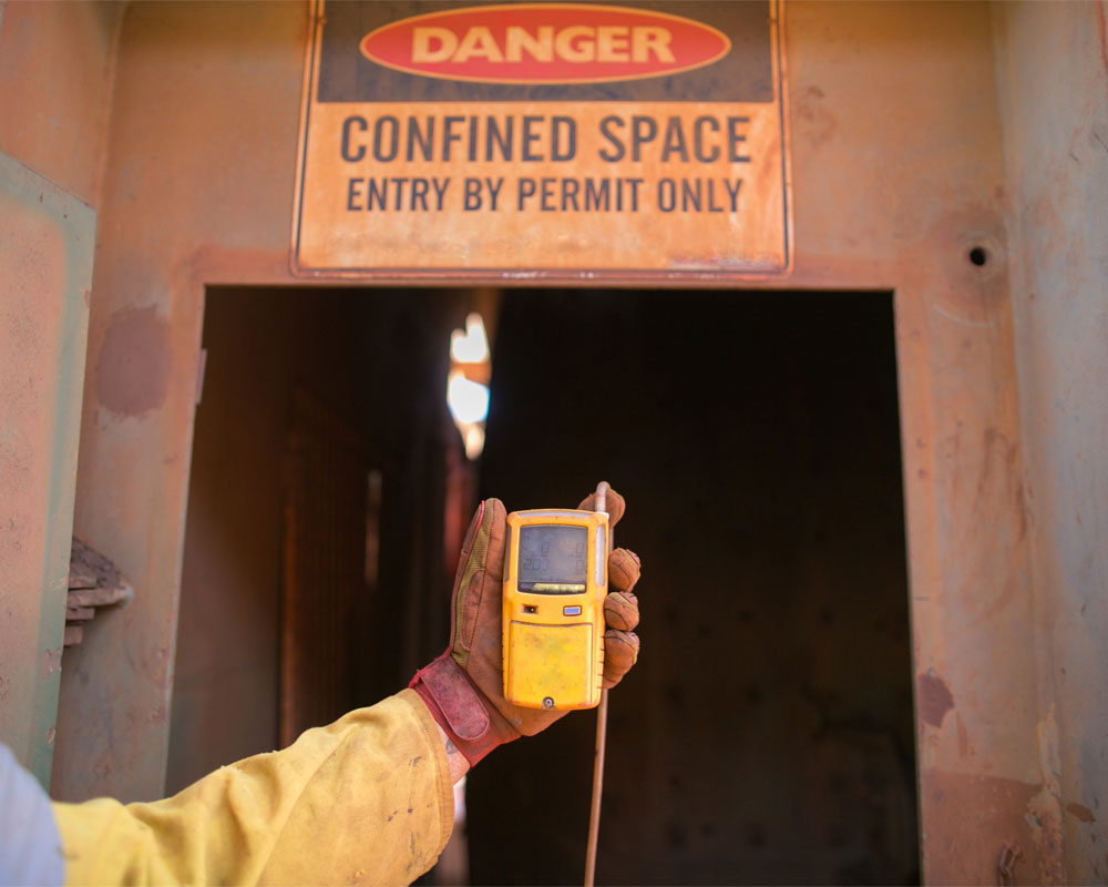 Hand holding a gas detector in front of a "Danger: Confined Space" sign, emphasizing safety compliance and hazard identification in workplace environments.
